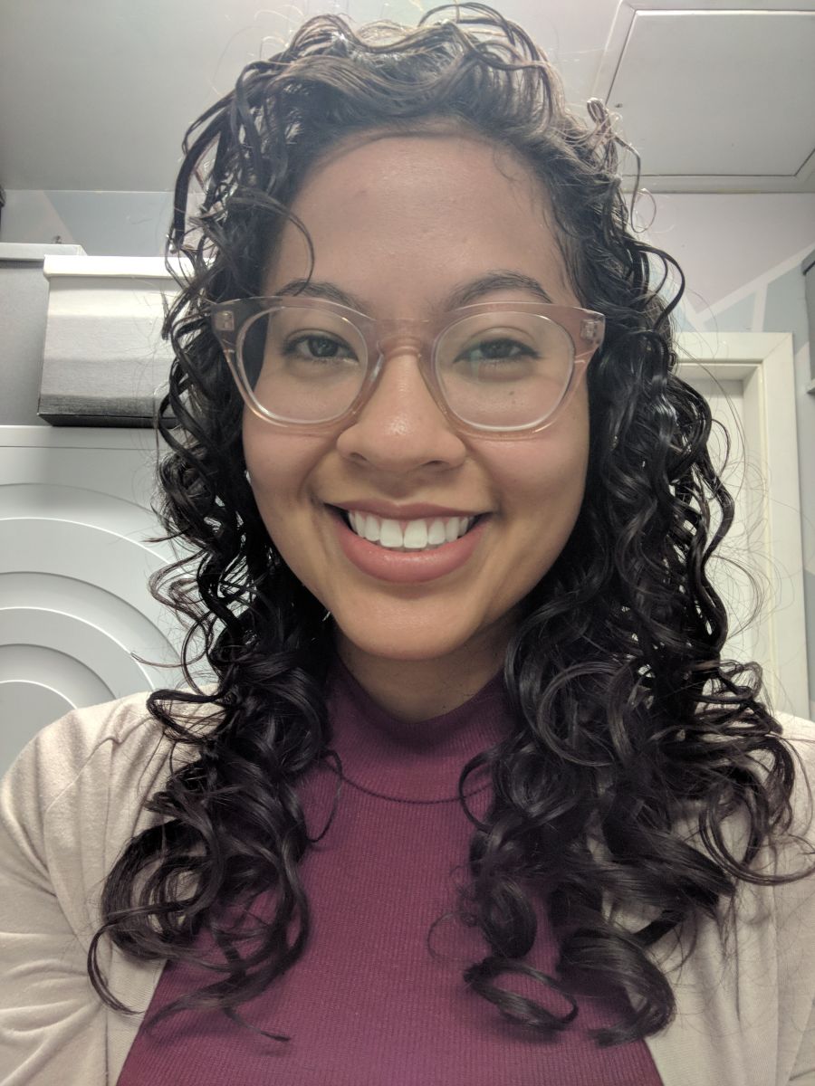 ieko is facing forward, smiling into the camera.  She has brown skin, curly hair, and is wearing pink glasses and a beige cardigan over a burgundy turtleneck. 