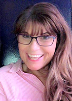 smiling photo of light skinned nonbinary woman with shoulder-length hair and glasses wearing pink blouse with collar