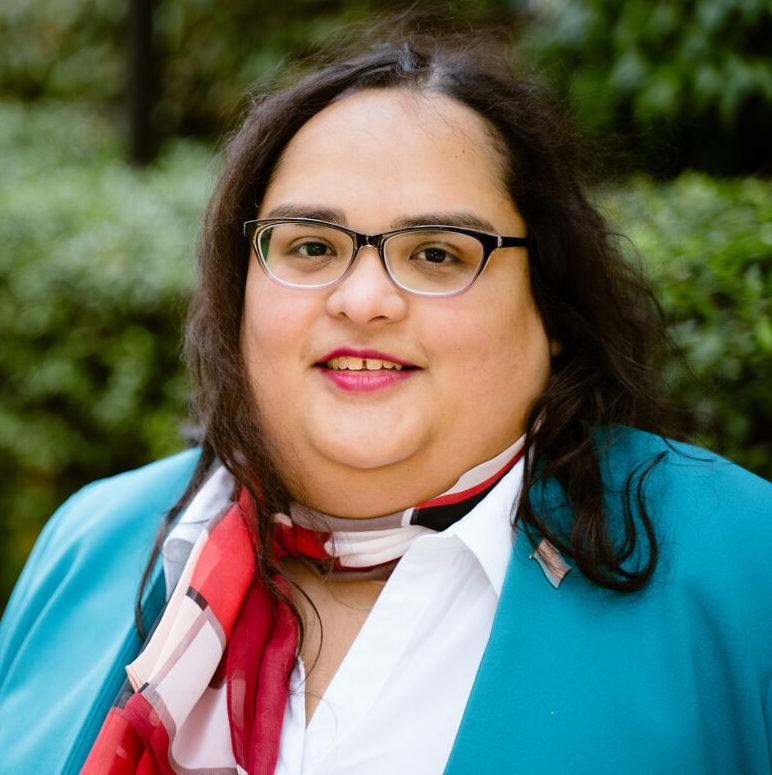 Latina trans woman with long brown wavy hair wearing glasses and turquoise blazer over white blouse with red scarf
